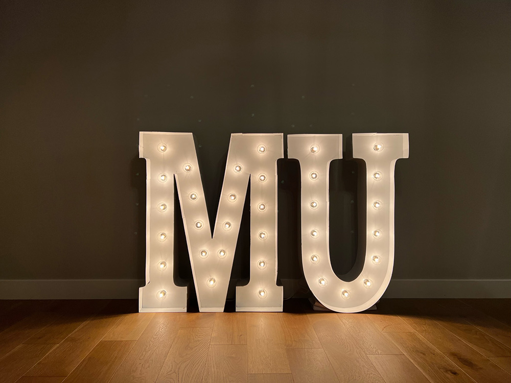 The acronym 'MU' in lit marquee letters with white bulbs