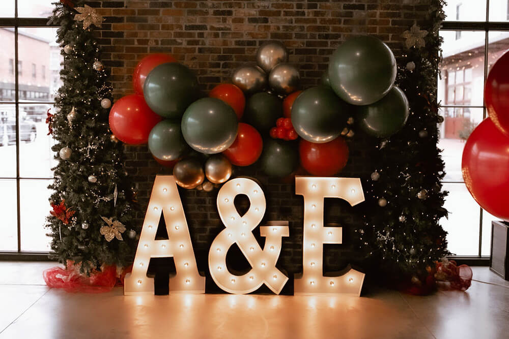 The initilals "A&E" in lit marquee letters with white bulbs with a black, red, green and gold balloon garland surrounded by two Christmas trees