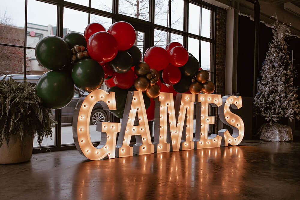 The word "Games" in lit marquee letters with white bulbs with a black, red, green and gold balloon garland