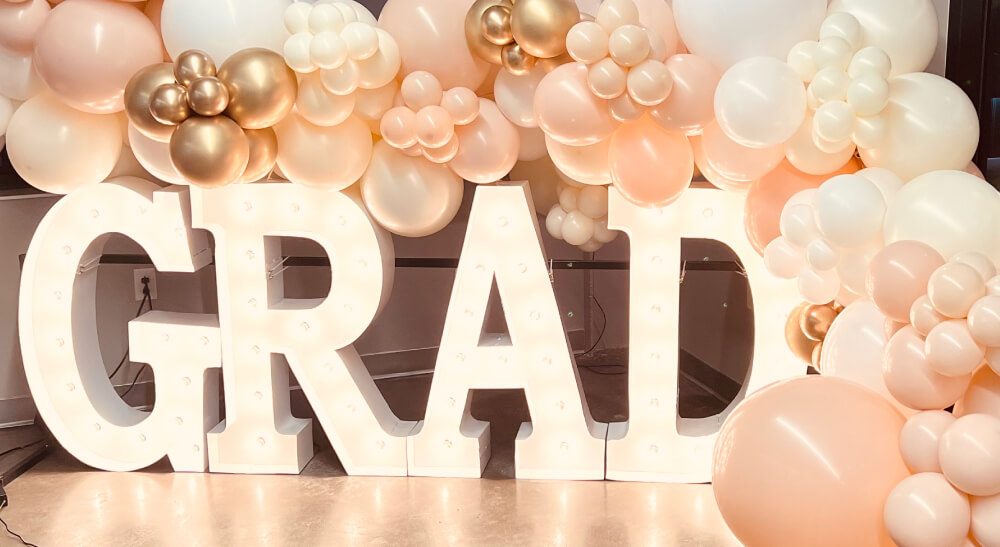 The word "Grad" in lit marquee letters with white bulbs with a peach, white, and gold balloon garland around the letters