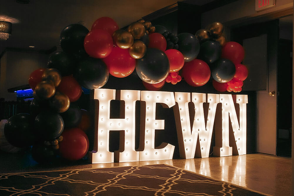 The word "Hewn" in lit marquee letters with white bulbs with a black, red, green, and gold balloon garland 