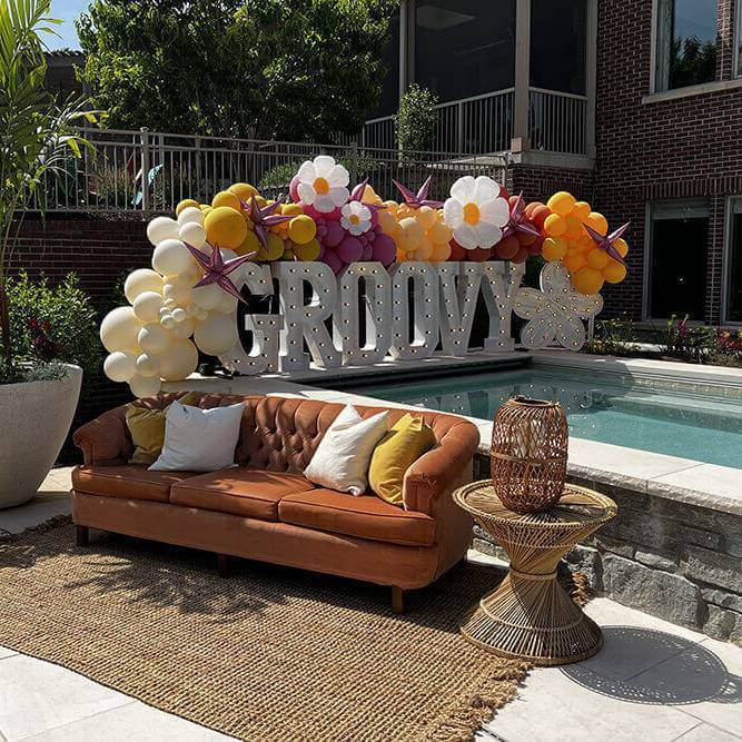 The word "Groovy" in marquee letters with a balloon garland next to an outdoor pool and a brown couch with a side table and a chair